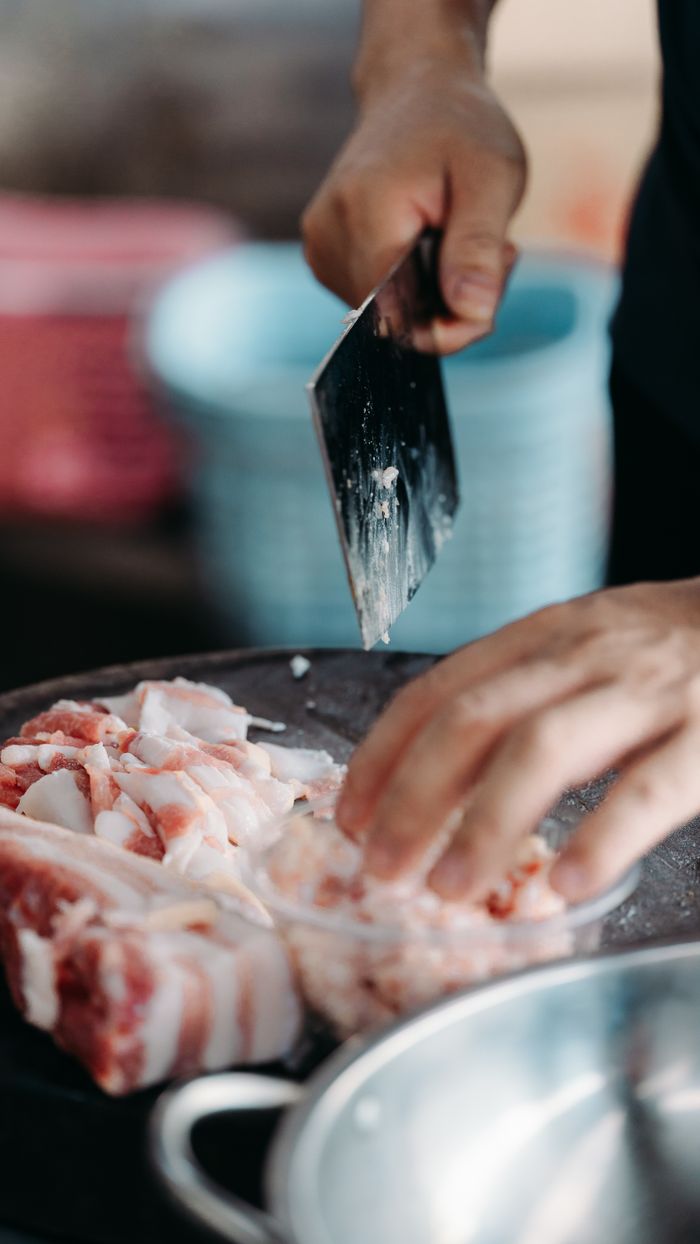 A chef skillfully slices fresh pork using a cleaver on a wooden board in a kitchen setting.